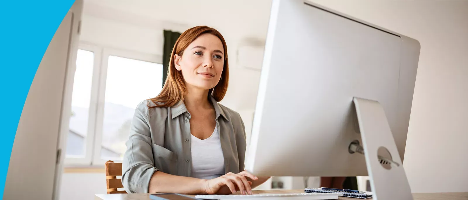 A woman smiling while working on a desktop computer in a bright home office.
