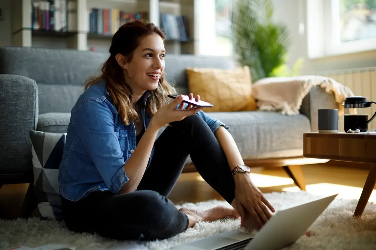 A smiling woman sits on a rug holding a phone toward her mouth with a laptop open nearby in a bright living room.