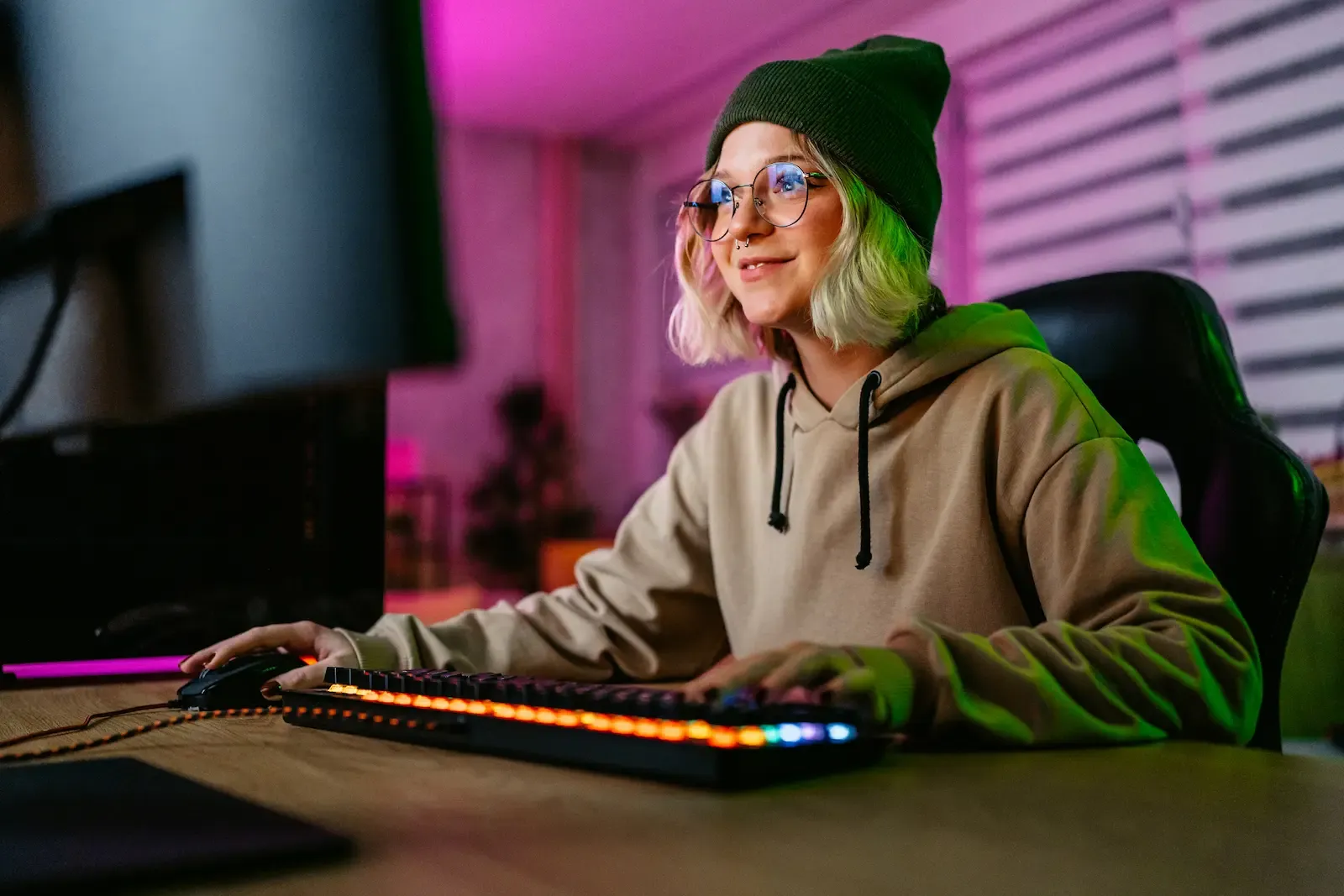 A young woman wearing a beanie and glasses smiles while using a backlit mechanical keyboard and mouse at a computer desk with colorful ambient lighting.