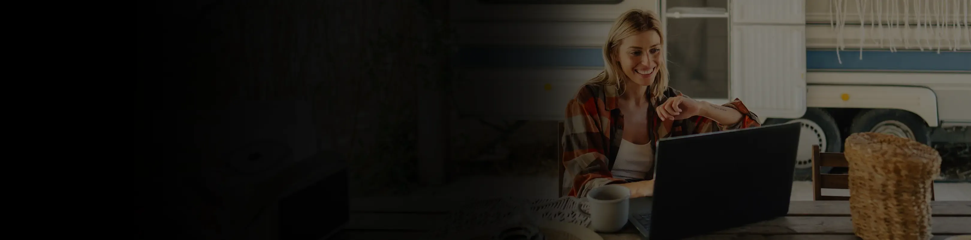 A smiling woman working on a laptop at a wooden table outside a vintage caravan trailer.