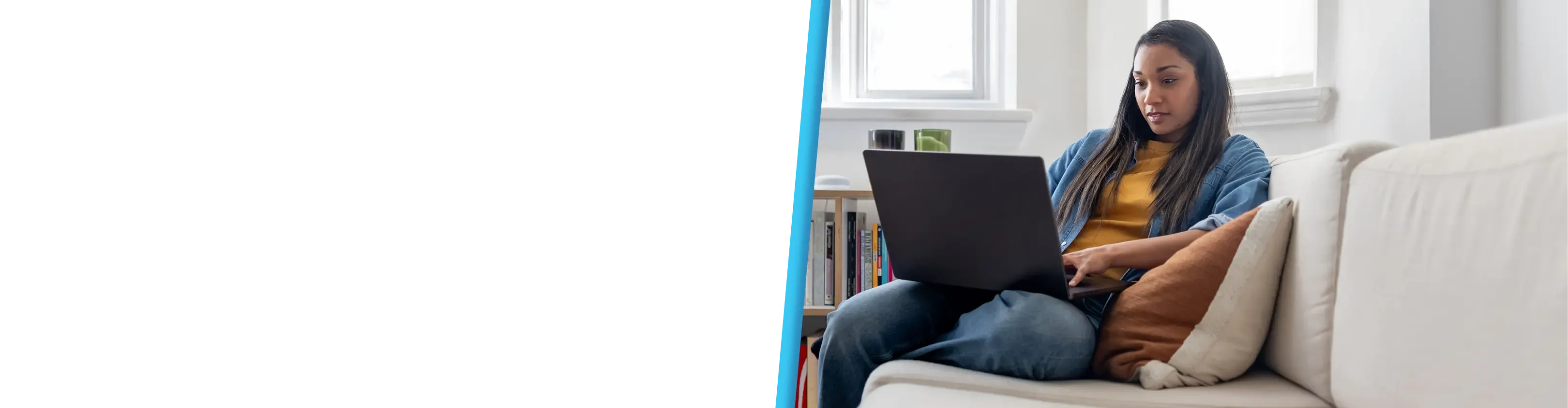 A woman in a yellow top and denim jacket types on a laptop while seated on a white couch.