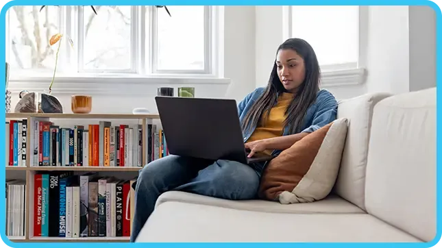 A woman in a yellow top and denim jacket types on a laptop while seated on a white couch.