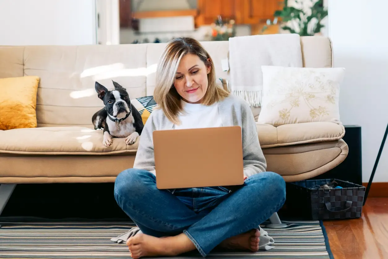 A woman sits cross-legged on the floor using a laptop while a Boston Terrier rests on the sofa behind her.