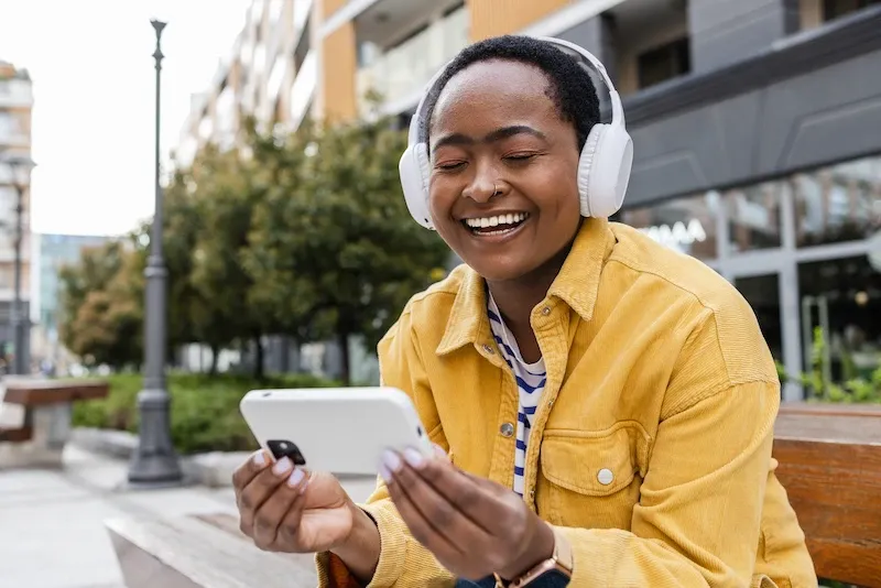 A happy woman in a yellow jacket wearing white headphones laughs while looking at her smartphone outdoors.