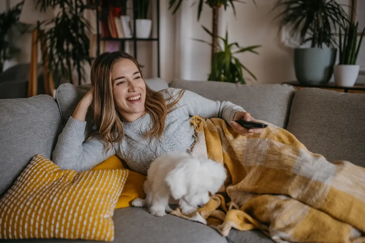 A laughing woman reclines on a gray sofa with a small white puppy and a yellow blanket, holding a remote control.