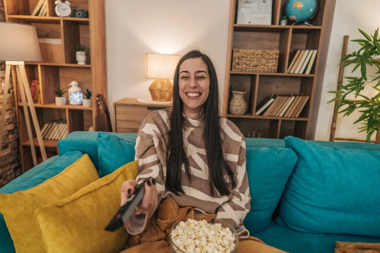 A laughing woman sits on a teal sofa with a bowl of popcorn and a remote control, enjoying a movie at home.