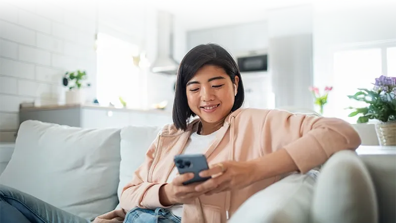 A smiling woman wearing a peach hoodie and wireless earbuds sits on a light-colored couch looking at her smartphone in a bright, modern living space.