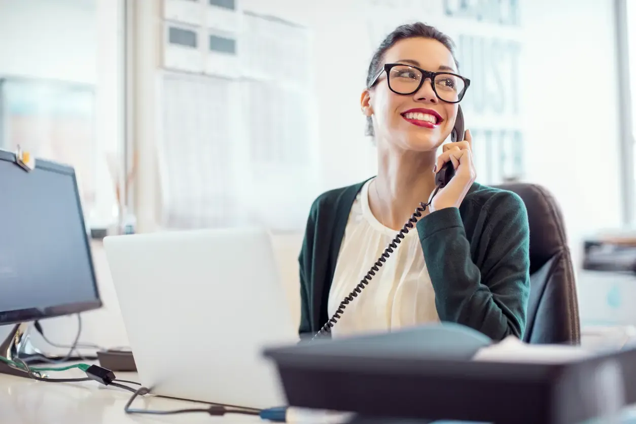 A smiling woman with glasses talks on a desk phone at her office workstation.