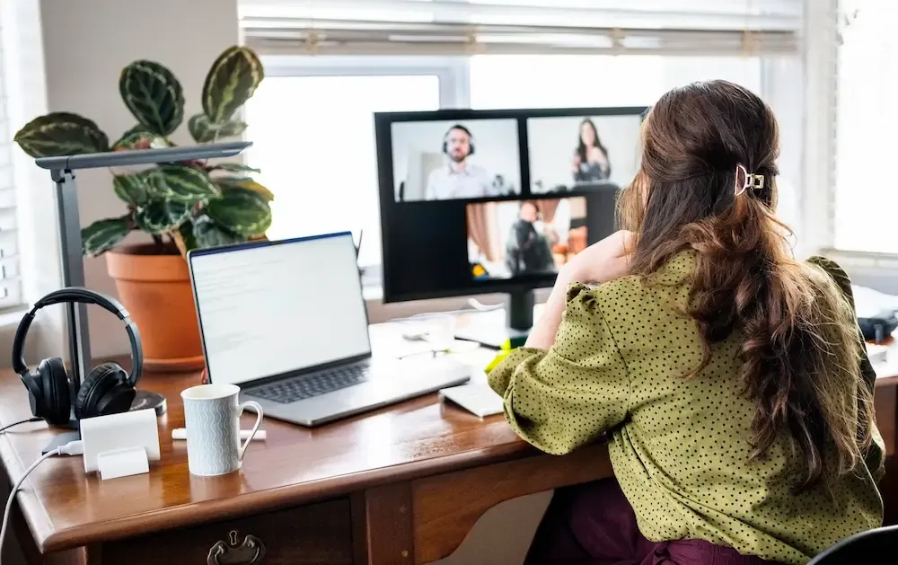 A woman viewed from behind sits at a wooden desk and participates in a video conference call displayed on a large computer monitor.