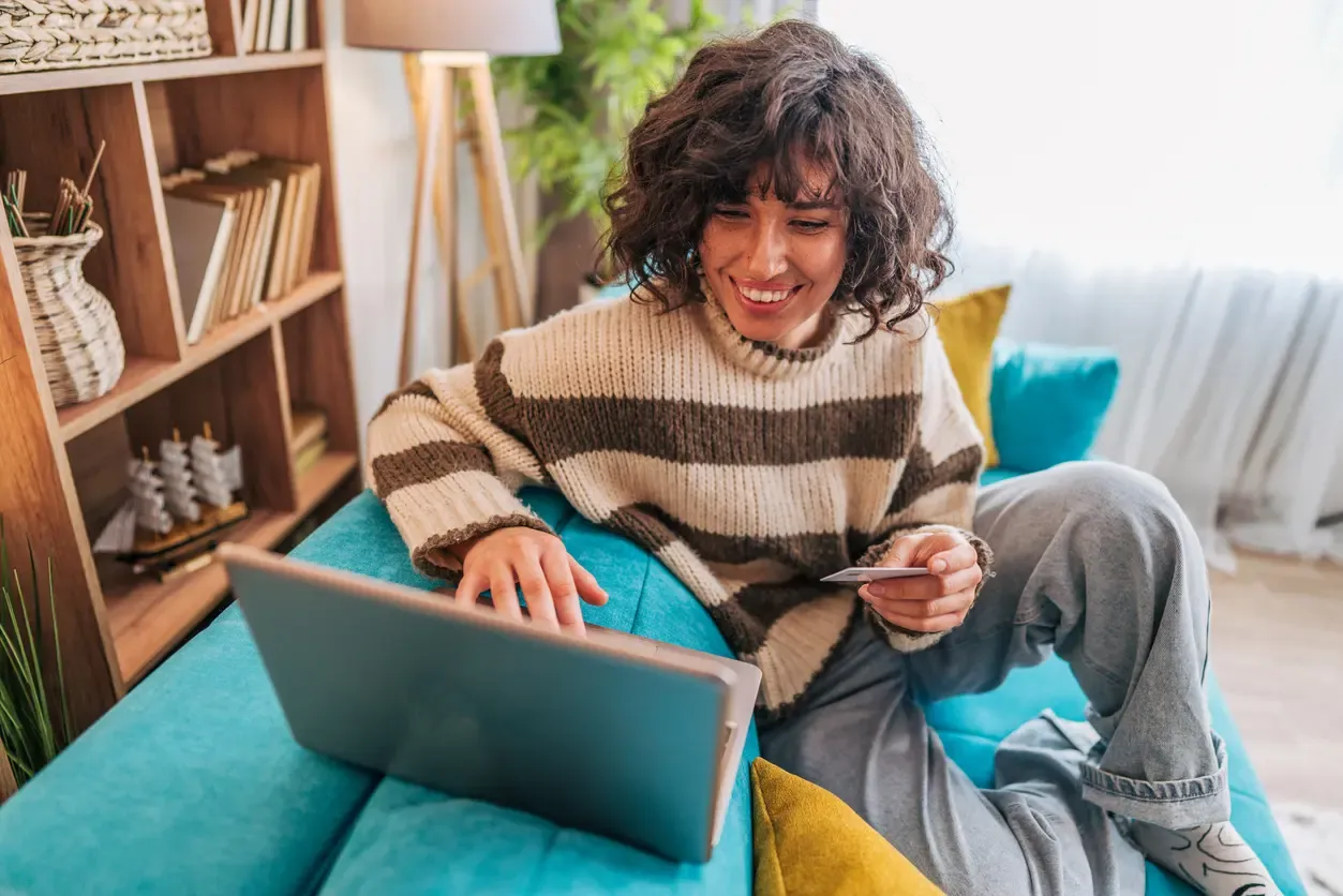 A smiling woman holds a credit card while shopping online on her laptop at home.