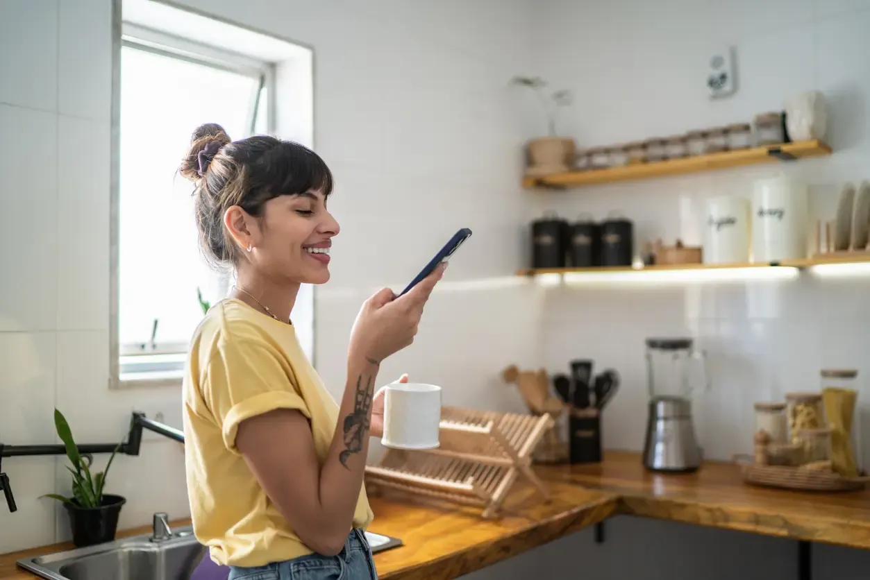 smiling woman in a yellow shirt holds a phone and mug in a bright modern kitchen.