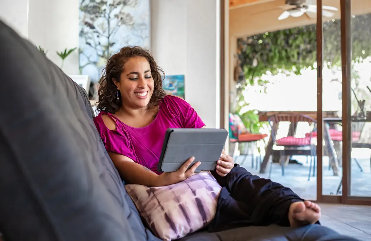 A smiling woman sitting on a gray couch looking at a tablet.