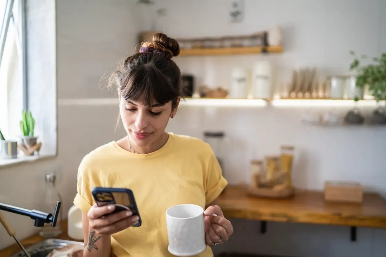A young woman checks her smartphone while holding a white coffee mug in her kitchen.