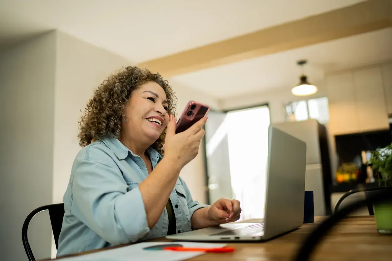 A smiling woman with curly hair holds her smartphone while working on a laptop in her kitchen.