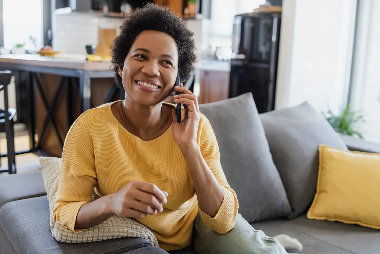 A smiling woman in a yellow sweater talks on a smartphone while sitting on a gray sofa at home.