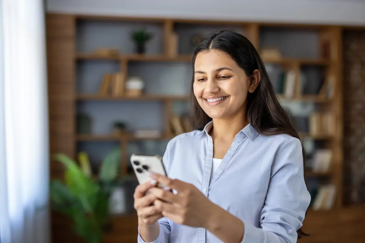 A smiling woman in a light blue shirt uses her smartphone in a home office.