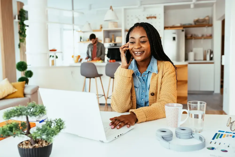 A smiling woman with braids talks on her smartphone while using a laptop at a white kitchen table.