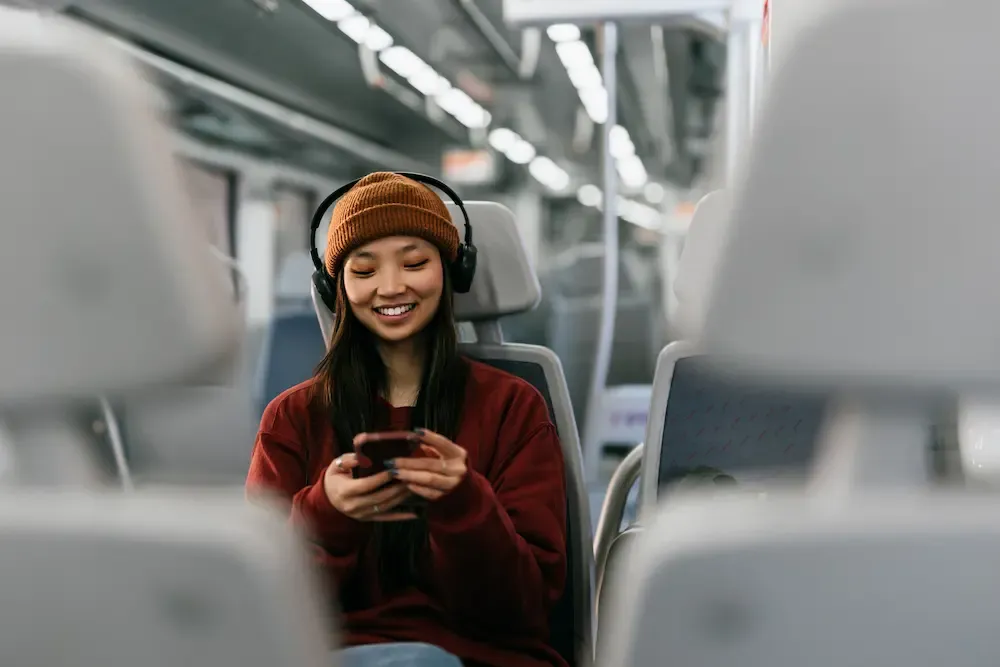 A smiling woman wearing a beanie and headphones looks at her smartphone while sitting on a train.