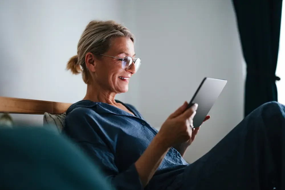 A smiling mature woman with glasses sitting in bed and looking at a digital tablet.