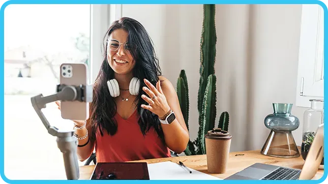 A smiling woman records herself on a phone gimbal at a desk with cacti.