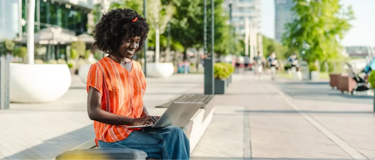 A smiling woman sits on a bench working on her laptop along a city plaza.