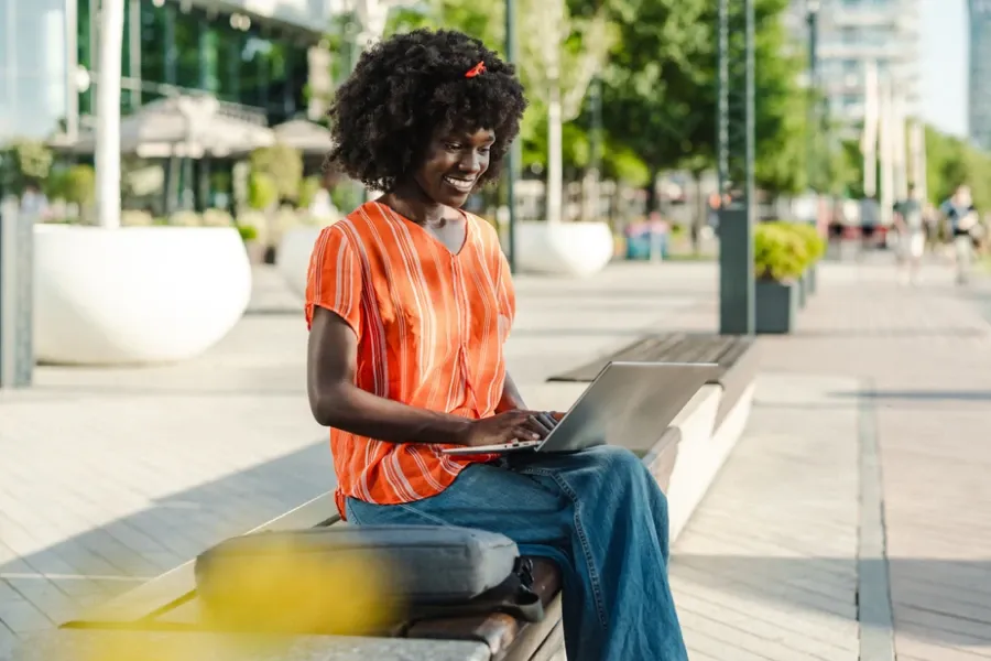 A smiling woman sits on a bench working on her laptop along a city plaza.