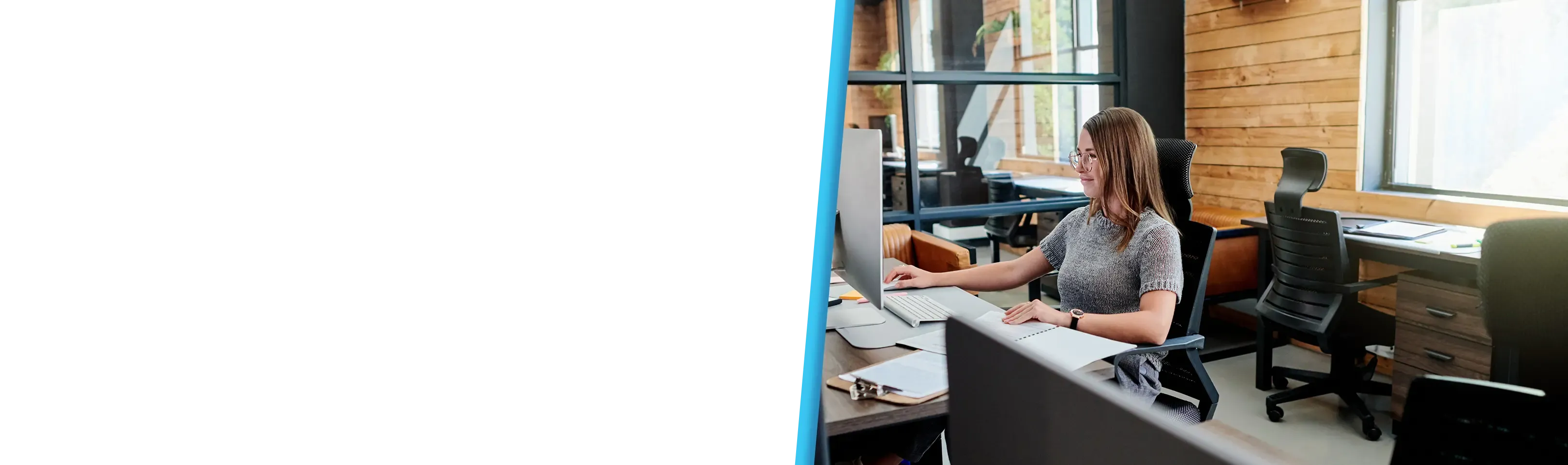 A woman wearing glasses works at a desktop computer in a bright, modern wood-accented office.