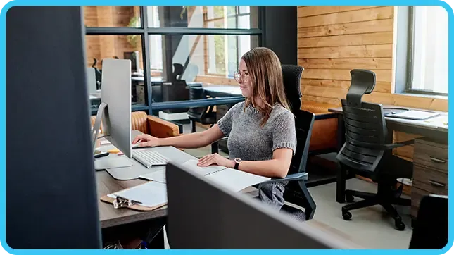 A woman wearing glasses works at a desktop computer in a bright, modern wood-accented office.