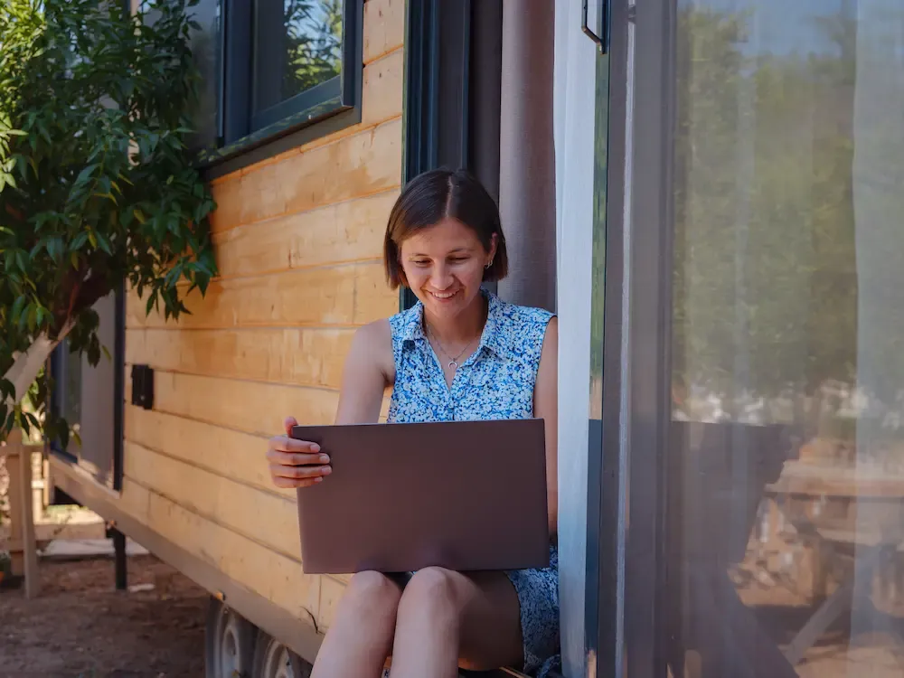 A smiling woman sits in the doorway of a wooden tiny house on wheels while working on a laptop.
