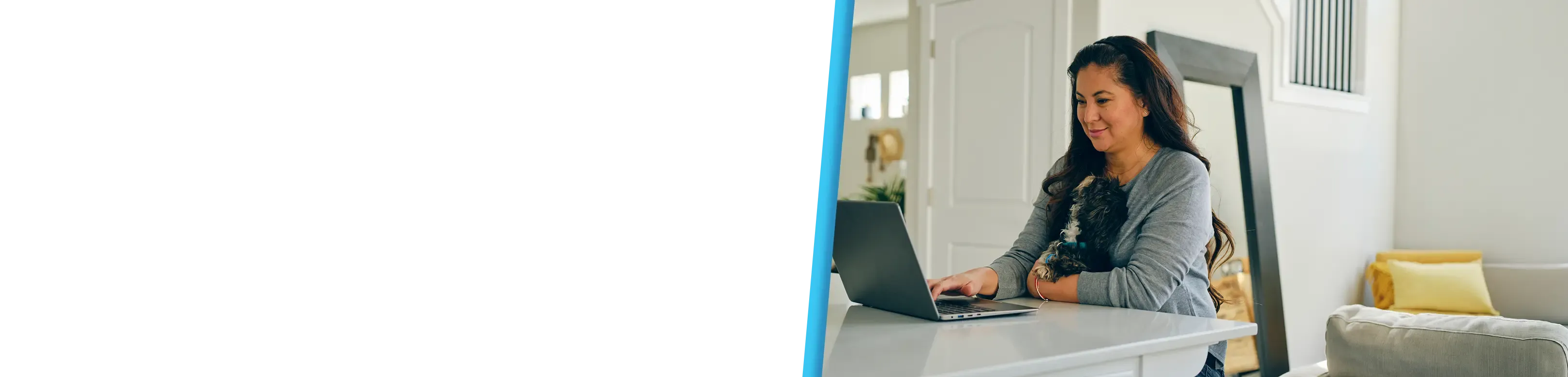 A woman sitting at a white kitchen island and working on a laptop while holding a small dog in her lap in a bright, modern home.