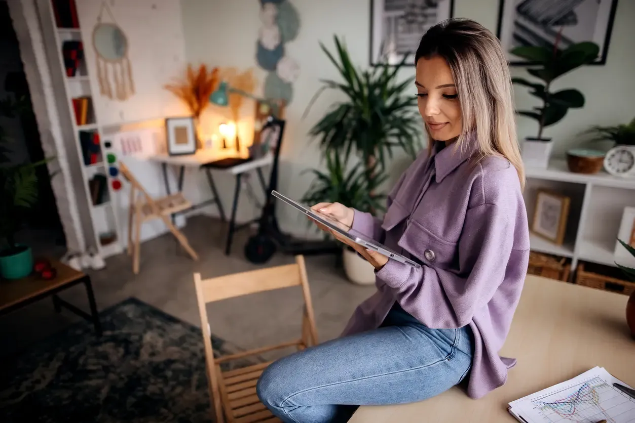 A woman in a purple jacket sits on a desk in a modern home office, focused on using a digital tablet.