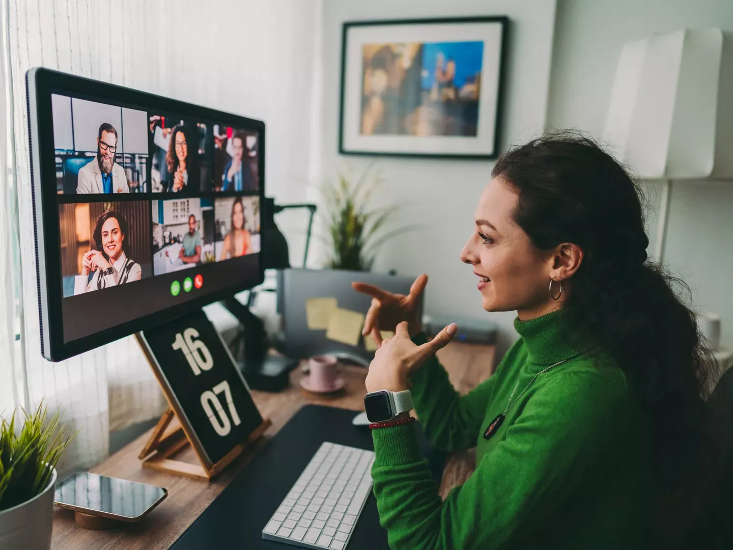 A woman in a green sweater uses hand gestures while participating in a group video call on a large computer monitor.