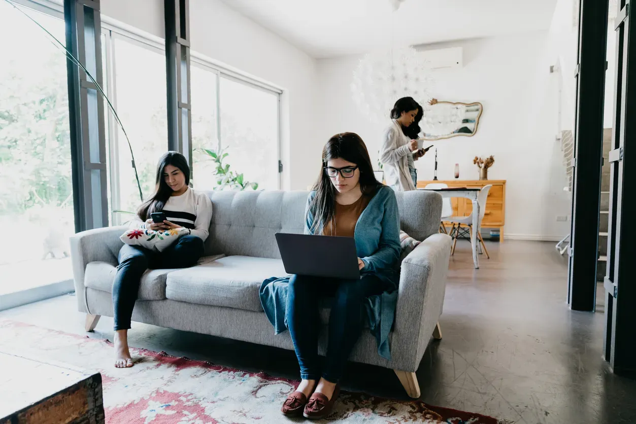 Two women sit on a couch, one using a laptop and one on her phone.