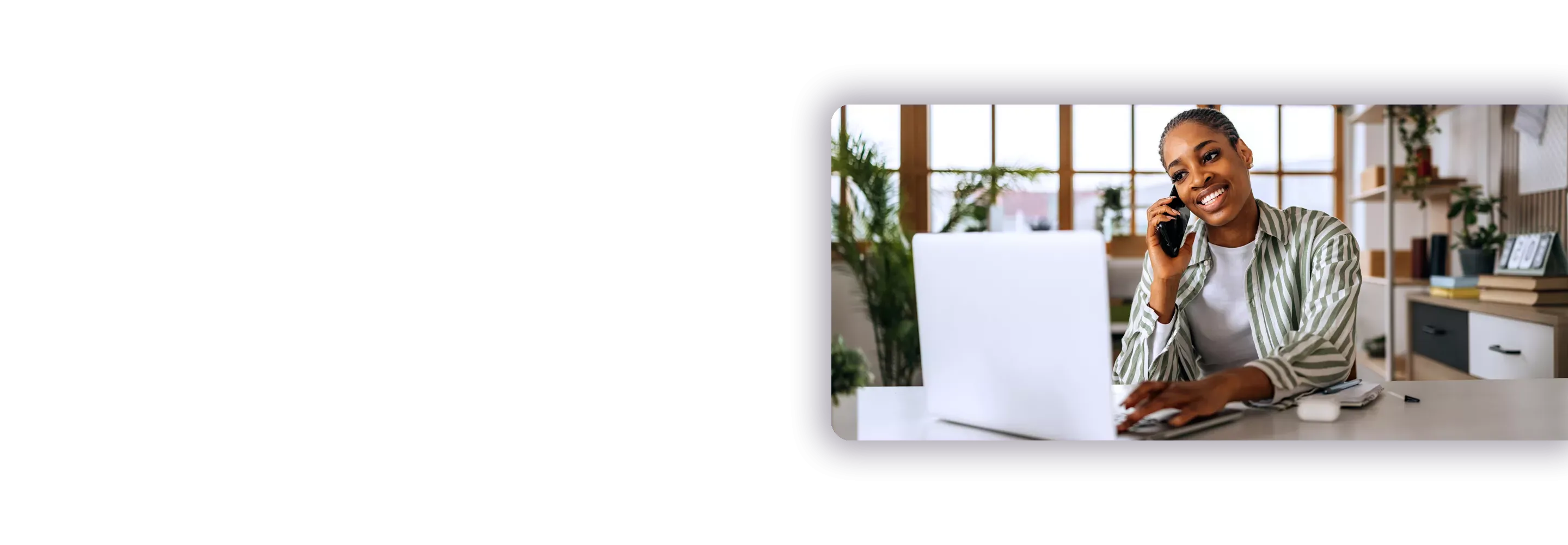 A smiling woman working on a laptop while talking on a mobile phone in a bright home office.