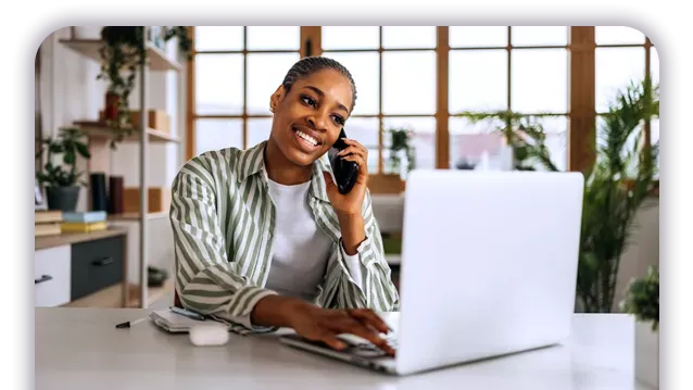 A smiling woman working on a laptop while talking on a mobile phone in a bright home office.