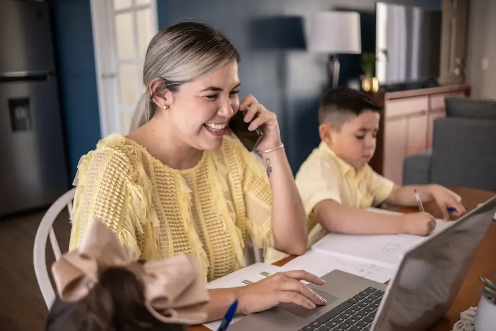 A laughing mother talks on her smartphone and works on a laptop while sitting at a table next to her young son who is focused on his schoolwork.