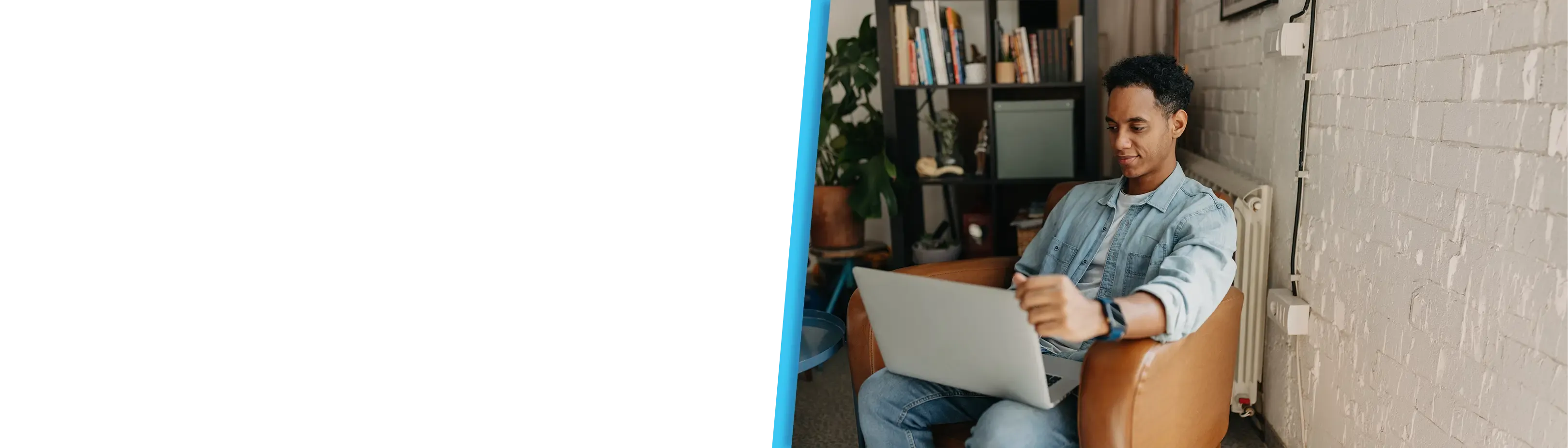 A young man in a denim shirt uses a laptop while seated in a brown leather chair at home.