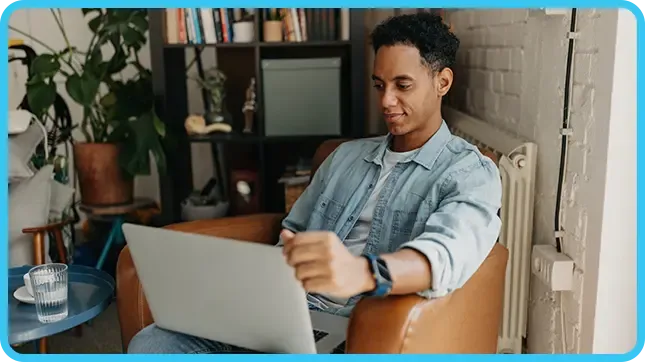 A young man in a denim shirt uses a laptop while seated in a brown leather chair at home.