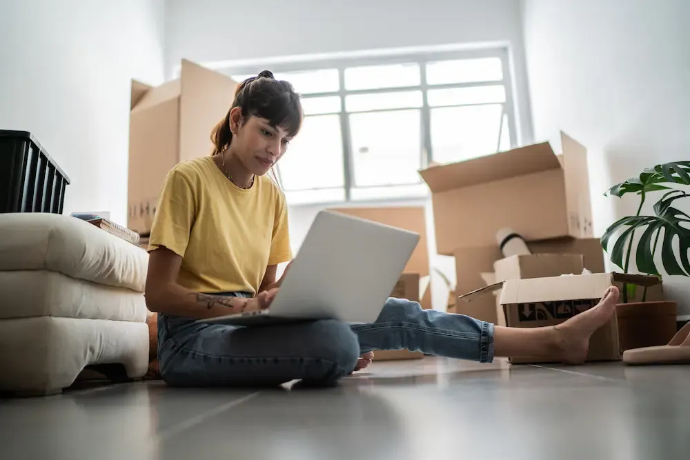 A young woman sitting on the floor of a new apartment using a laptop surrounded by moving boxes.