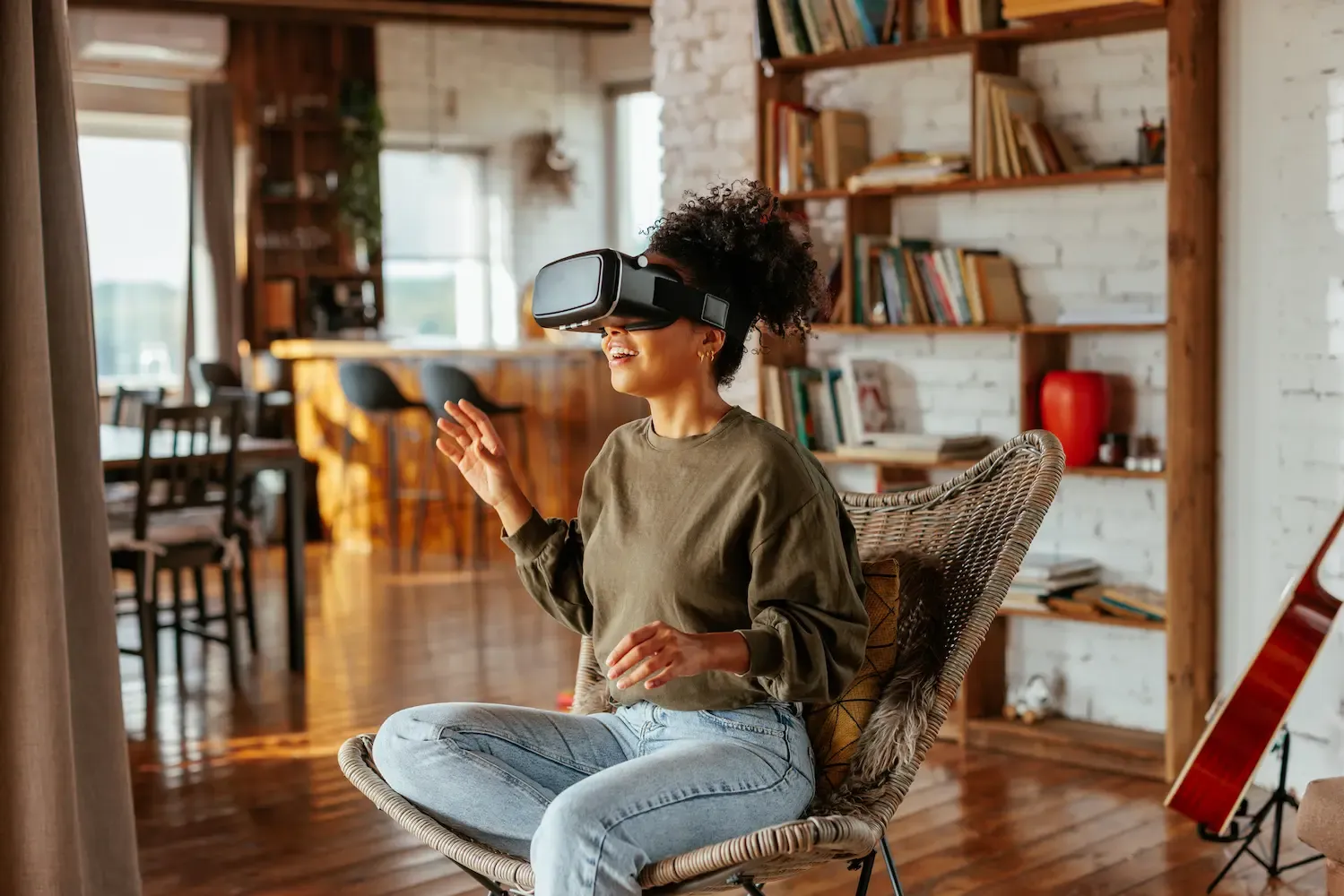 A young woman wearing a virtual reality headset sits in a wicker chair in a modern, sunlit living room with her hands raised in engagement.